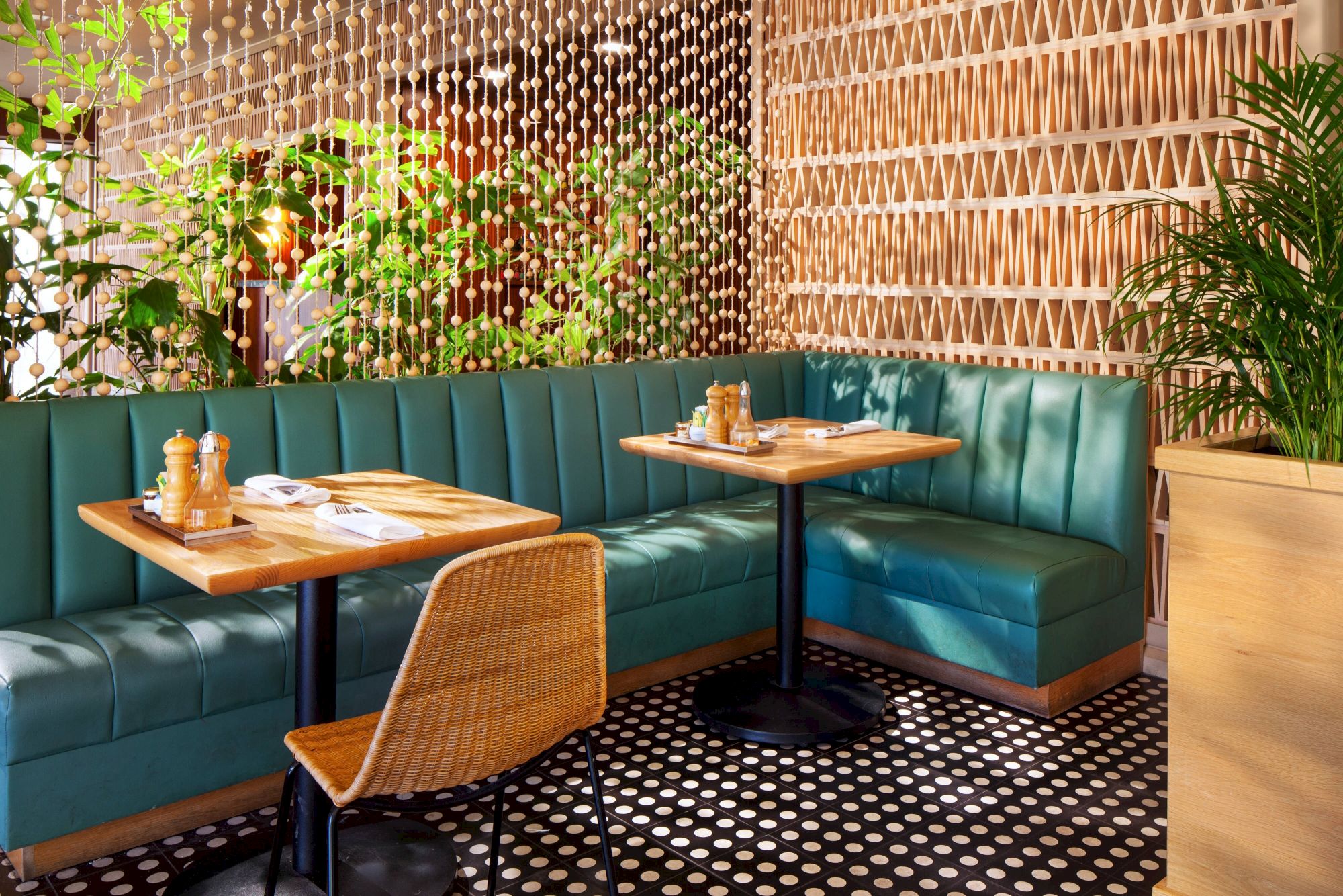 A cozy restaurant area with teal booth seating, wooden tables, a woven chair, vibrant plants, and patterned flooring.