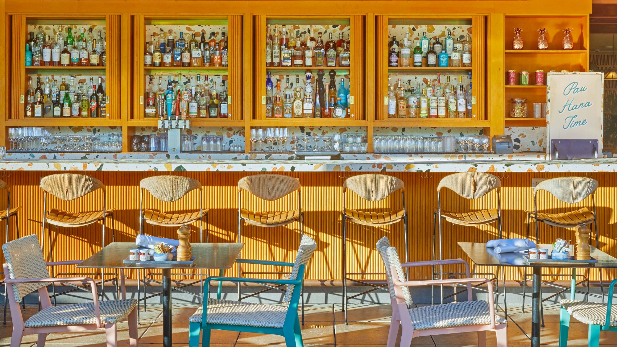 The image shows a brightly lit bar setup with a variety of bottles on shelves, chairs at the counter, and several tables with seating.