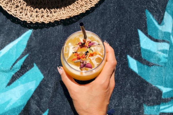 A hand holding a decorated drink with a straw, next to a straw hat on a tropical-themed background.