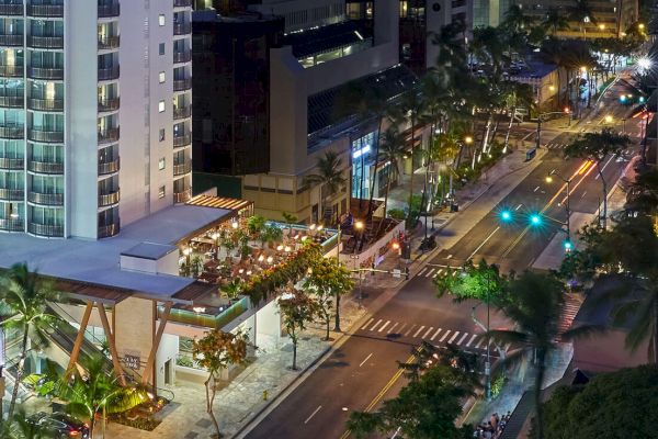 An aerial view of a lively city street at night, lined with palm trees and illuminated buildings, featuring a bustling outdoor restaurant.