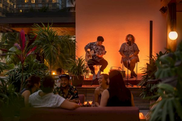 A cozy nighttime scene with live musicians performing outdoors; people are enjoying the ambiance surrounded by plants and soft lighting.