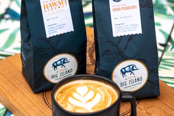 A latte with latte art in a black cup sits on a wooden board with two bags of coffee, featuring a leafy background.
