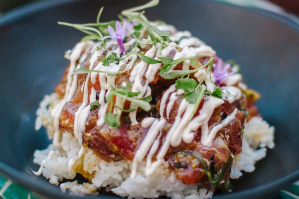 A dish of rice topped with a colorful mix of diced ingredients, drizzled with sauce, and garnished with sprouts and flowers.