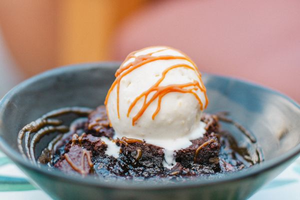 A black bowl with a chocolate dessert topped with vanilla ice cream and caramel drizzle, placed on a patterned table.