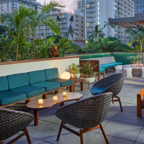 The image shows an outdoor patio with stylish seating, plants, and a fire pit, surrounded by urban high-rise buildings in the background.