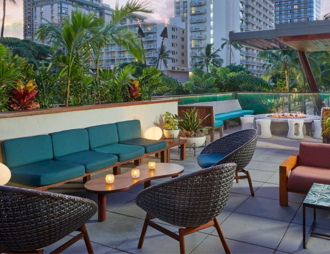 The image shows an outdoor patio with stylish seating, plants, and a fire pit, surrounded by urban high-rise buildings in the background.