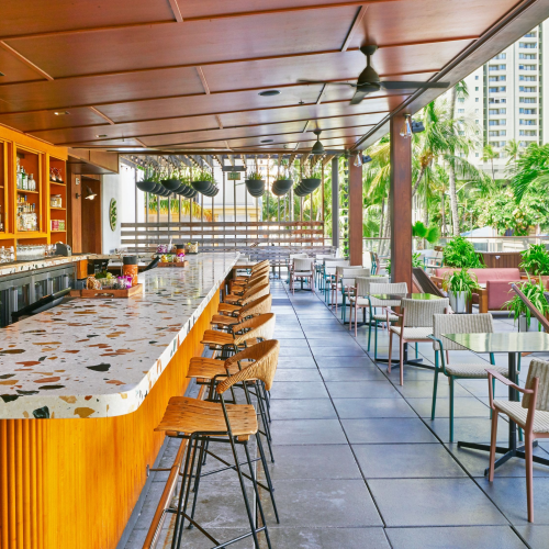 An outdoor bar with wooden decor, terrazzo countertops, and seating, surrounded by greenery and tall buildings in the background.