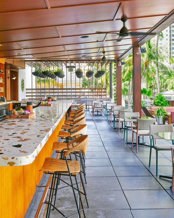 An outdoor bar with wooden decor, terrazzo countertops, and seating, surrounded by greenery and tall buildings in the background.