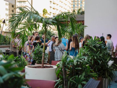 A group of people socializing at an outdoor event surrounded by lush green plants and a city backdrop.