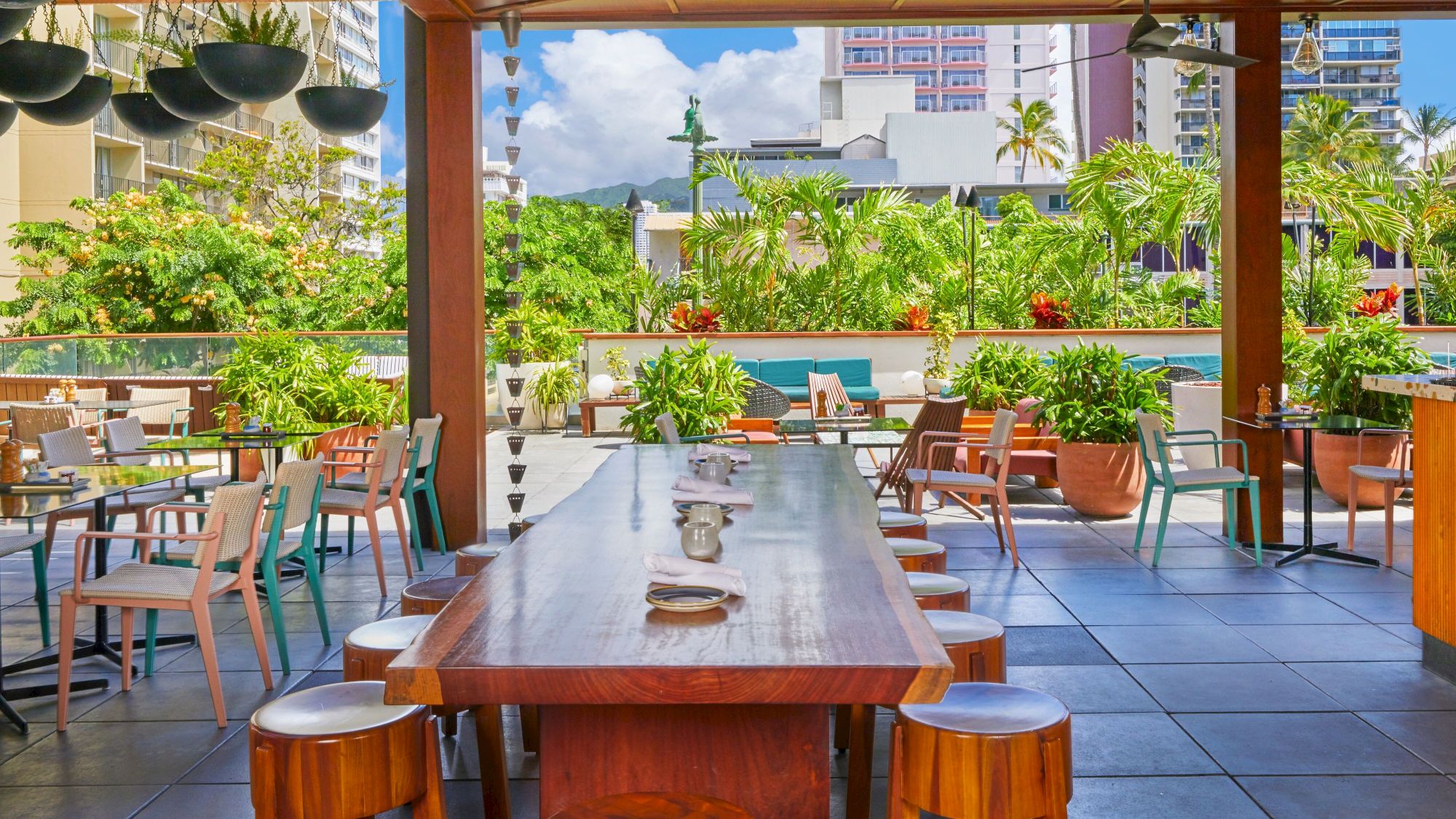 The image shows an outdoor restaurant patio with wooden tables and chairs, surrounded by greenery and city buildings in the background.