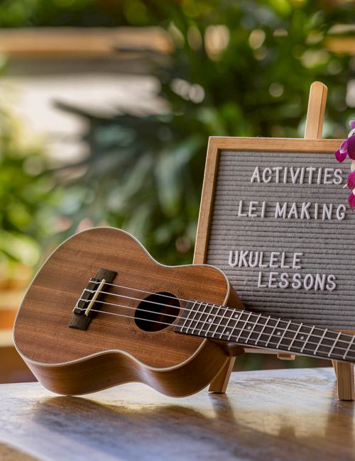 A ukulele rests on a table beside a sign listing activities like lei making and ukulele lessons, adorned with an orchid lei.