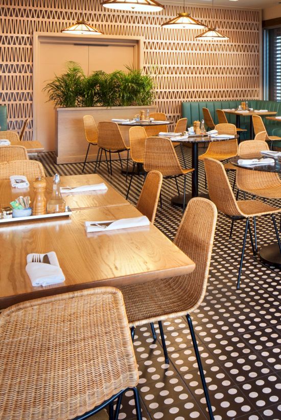 The image shows a modern restaurant interior with wooden tables, wicker chairs, green booth seating, and a geometric tile floor.