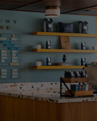 A coffee shop interior with a menu board, espresso machine, shelves with cups and coffee bags on a light blue wall.