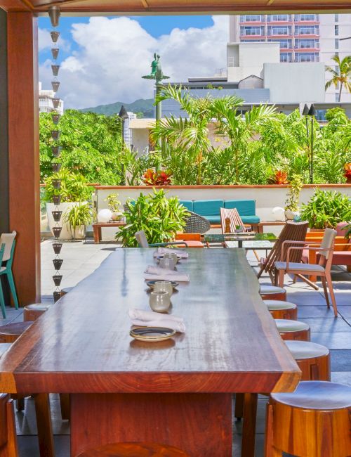 An outdoor patio area with wooden tables and chairs, surrounded by lush greenery and city buildings in the background.