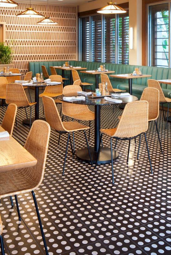 A modern restaurant interior with wooden tables, wicker chairs, green booth seating, patterned walls, and a checkered floor pattern.