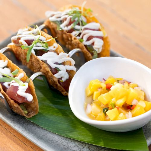 The image shows three tacos with sauce drizzled on top and a side of mango salsa in a small bowl, served on a platter with a leaf.