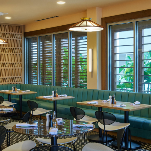 A modern restaurant interior with teal booth seating, black chairs, pendant lighting, and windows showing greenery outside.