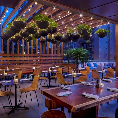 An outdoor dining area with hanging plants, string lights, and wooden tables set for guests, creating a cozy evening ambiance.