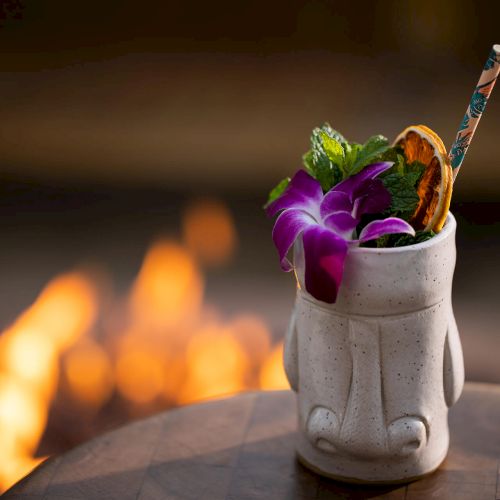 A tiki mug filled with a colorful cocktail, garnished with flowers and greenery, is placed on a table against a blurred fiery background.