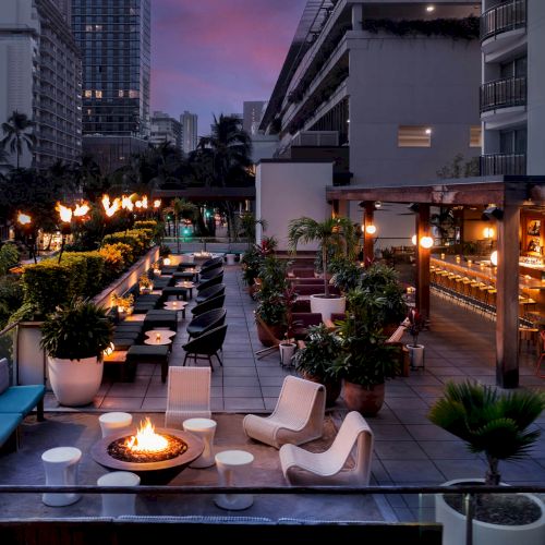 A rooftop lounge with comfy seating, a fire pit, outdoor bar, and cityscape views at sunset, creating a cozy and inviting atmosphere.