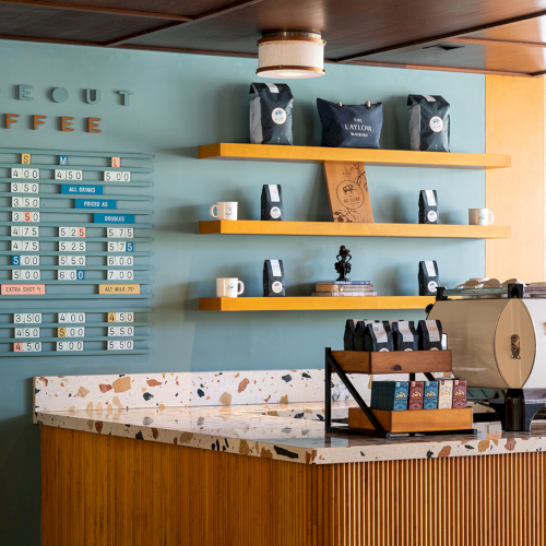 The image shows a coffee shop with a menu board, shelves of bags and cups, and a coffee machine on a marble counter.