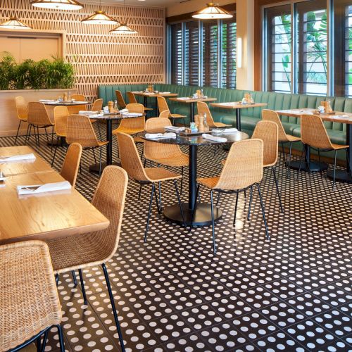 A modern restaurant interior with wooden tables, wicker chairs, patterned floor, and green cushioned seating, well-lit and inviting.