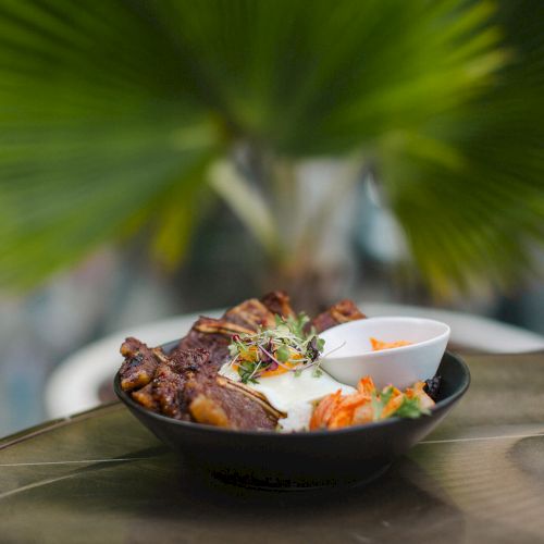 A bowl of grilled meat with vegetables, rice, and a small sauce dish is placed on a table, with a blurred green plant in the background.
