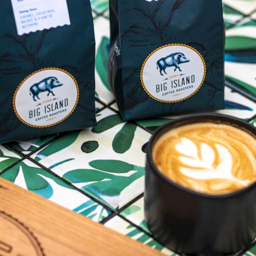 Two coffee bags labeled "Big Island" are next to a latte in a black mug with latte art on top, set on a leafy patterned surface.