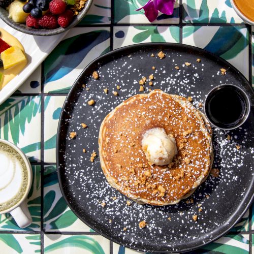 A table with pancakes topped with butter, a small syrup container, assorted fruit, a latte, and orange juice on patterned tiles.