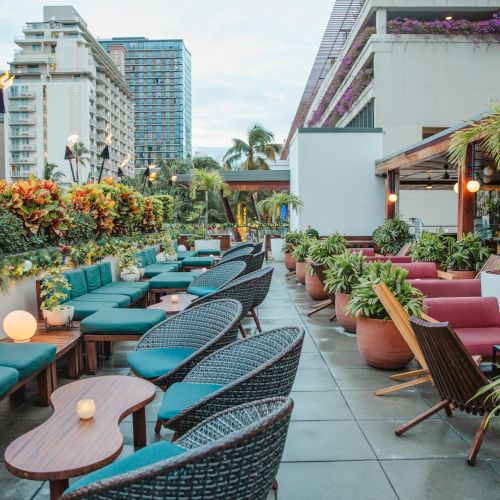 The image shows a stylish outdoor rooftop patio with cozy seating, tables, and lush plants, set in an urban setting with tall buildings.