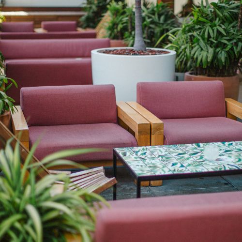The image shows a seating area with red cushioned chairs, a tiled table, and green plants around, creating a cozy and inviting atmosphere.