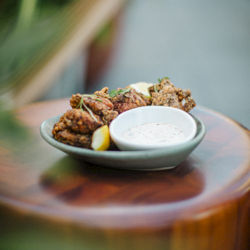 A plate with fried chicken pieces and a lemon wedge, accompanied by a bowl of dipping sauce, is placed on a wooden surface.