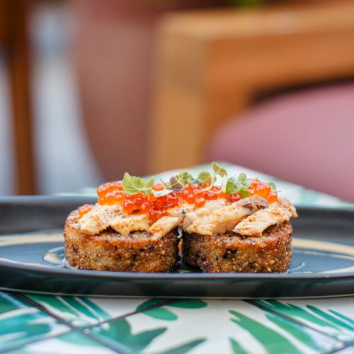 The image shows two slices of sushi or crispy rice topped with fish, red caviar, and microgreens on a black plate.