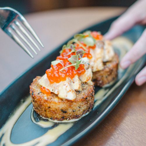 Two gourmet crab cakes topped with cream and red garnish on a black platter, with a fork and hand nearby, set on a wooden table.