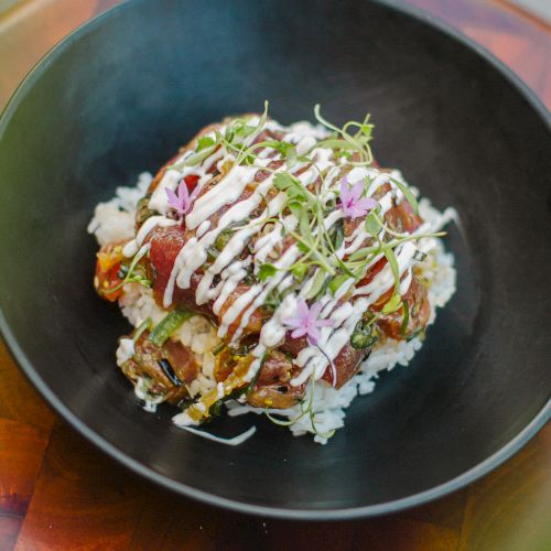 A bowl of rice topped with diced tuna, drizzled sauce, and garnished with sprouts and edible flowers is displayed on a wooden surface.