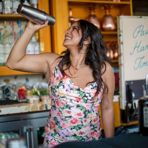 A woman in a floral dress is smiling and shaking a cocktail shaker behind a bar, with shelves of bottles and decor in the background.