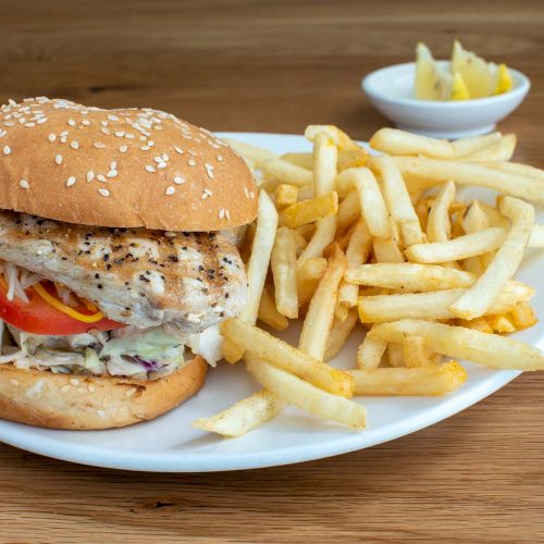 A grilled chicken sandwich with lettuce and tomato, served with a side of French fries on a white plate, with pickles in a small dish.