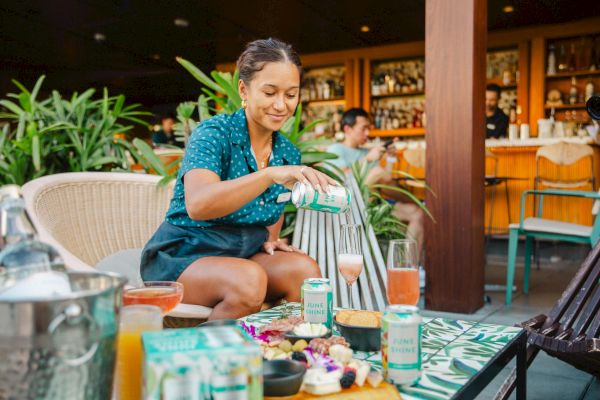 A person is pouring a drink at an outdoor table with snacks and drinks, surrounded by greenery and a bar in the background.